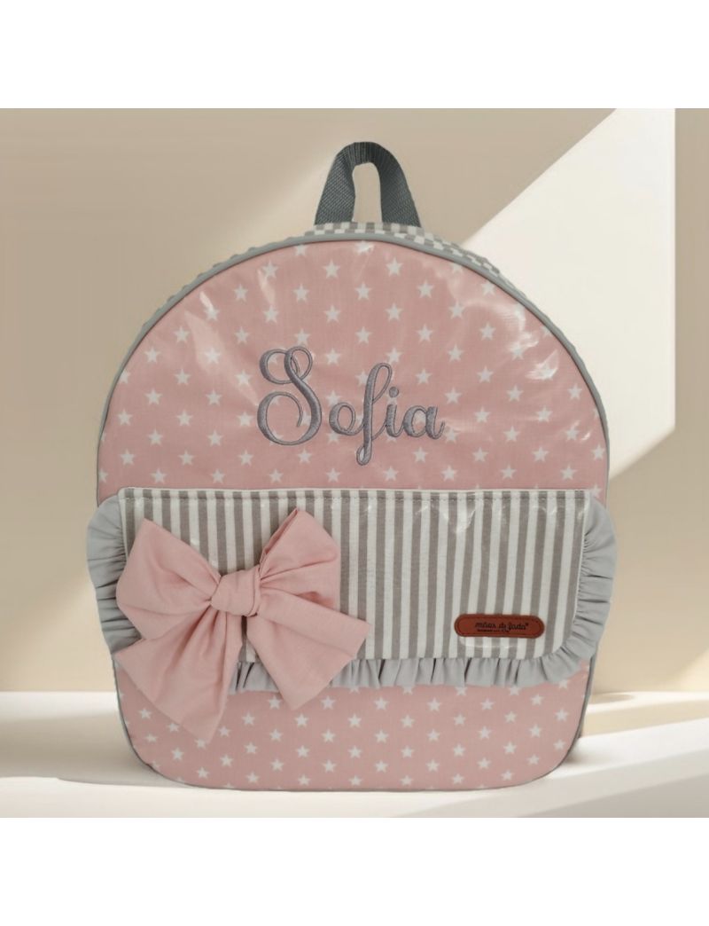 Mochila de criança com fundo rosa e estrelas brancas, conjugadas com riscas brancas e cinzentas. Bolsa frontal com pala com riscas cinzas e brancas, folho cincento e laço cor-de-rosa. Nome Sofia bordao a cinza na parte superior da mochila (tecido rosa com