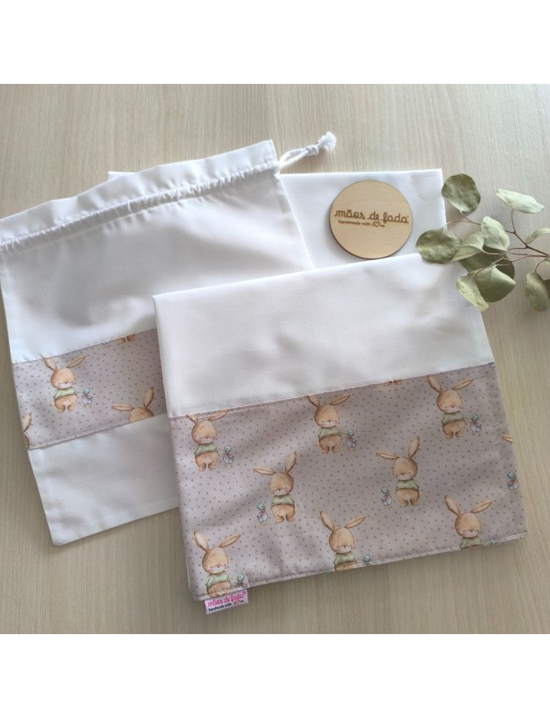 Lençol de cima, lençol de baixo e saquinho. O lençol de cima e o saquinho possuem uma barra com padrão de coelhinhos amorosos em tons de bege e castanho 