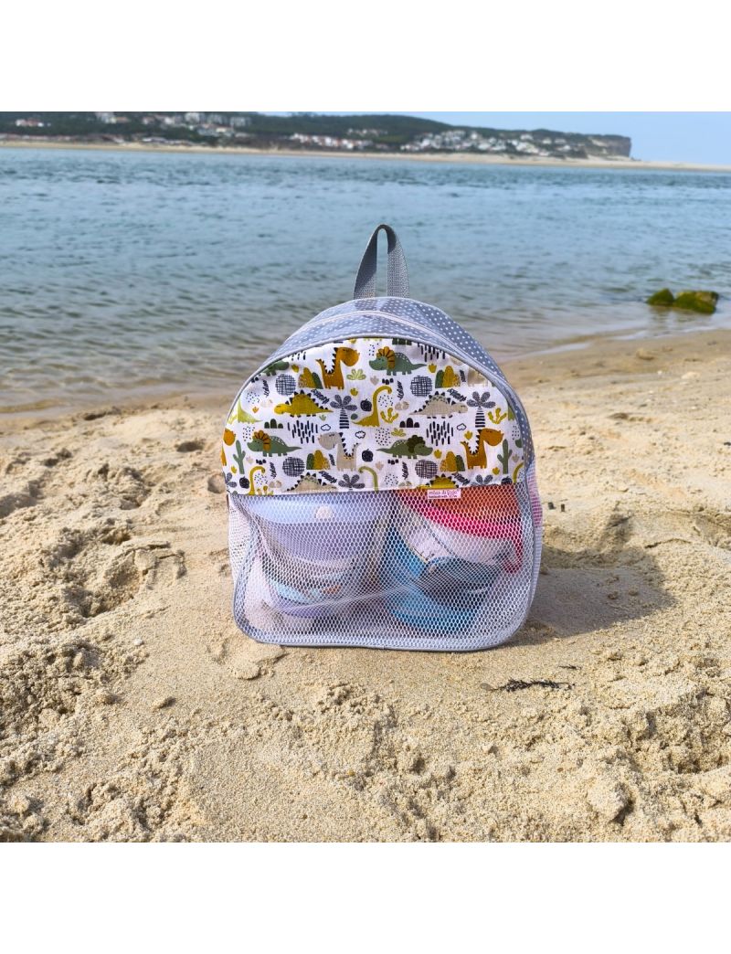 Mochila de praia com padrão de dinossauros em tons de verde, amarelo torrado e cinzento, conjugado com tecido cinzento com bolas brancas. Rede branca e alças cinzentas