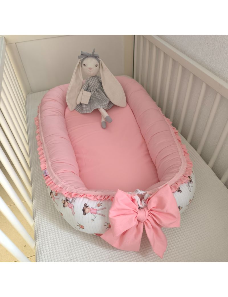 Ninho redutor de berço versátil para bebé, em tecido algodão respirável com padrão de bonequinhas no exterior e interior em rosa liso. Laço frontal e folhinho rosa a contornar o ninho. Ninho colocado dentro do berço  branco  