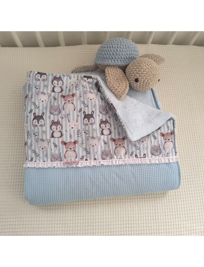 Manta de bebé em tecido waffle azul e estampado de animais fofos sobre riscas azuis e brancas. A manta tem um folho branco de detalhe e o interior é forrado a tecido coralina azul-claro. Está dobrada em cima de uma caminha, com uma tartaruga em amigurumi