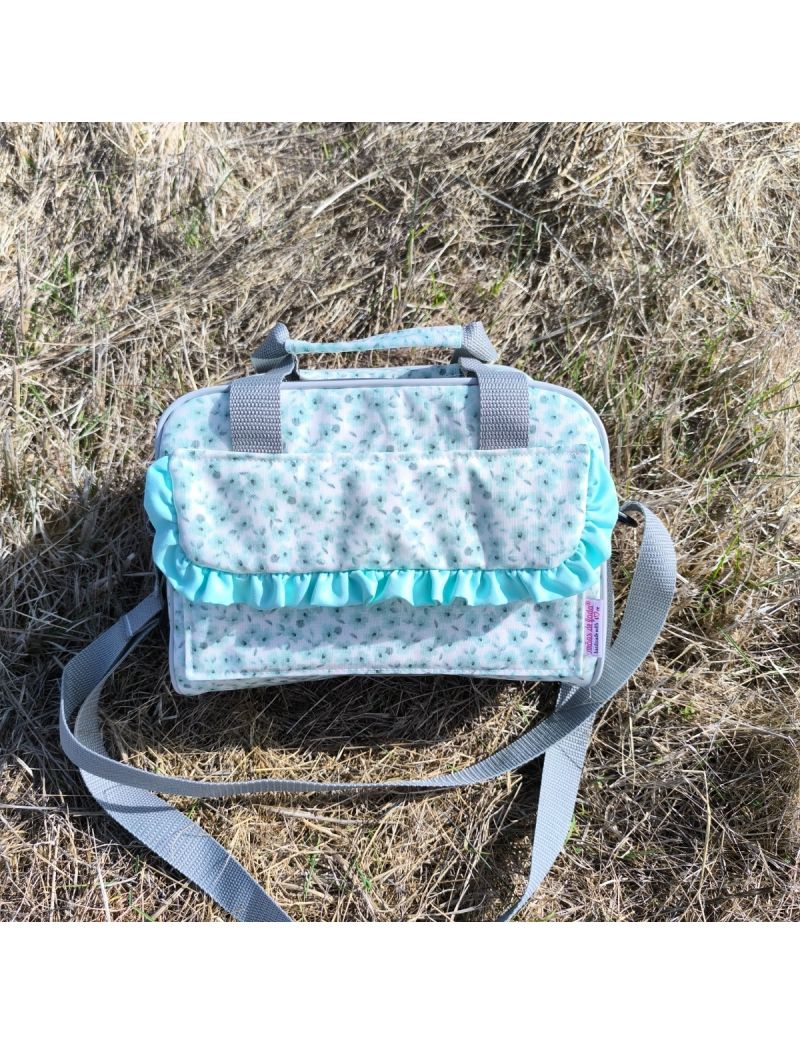 Lancheira XS com alça ajustável e alça de mão cinzenta, com padrão de pequenas flores verdes, e detalhe de folho em verde na pala frontal, colocada sobre relva seca ao ar livre