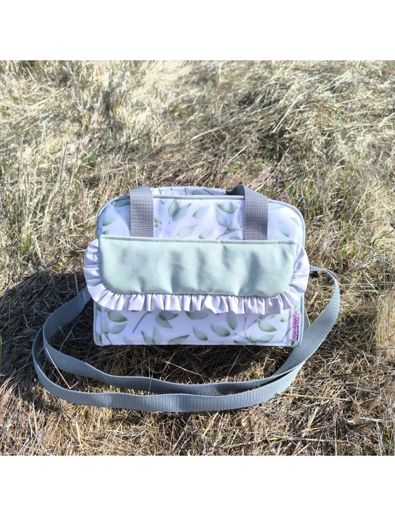 Lancheira XS com alça ajustável e alça de mão cinzenta, com padrão floral em tons de verde e detalhe de folho em cizento na pala frontal, colocada sobre relva seca ao ar livre