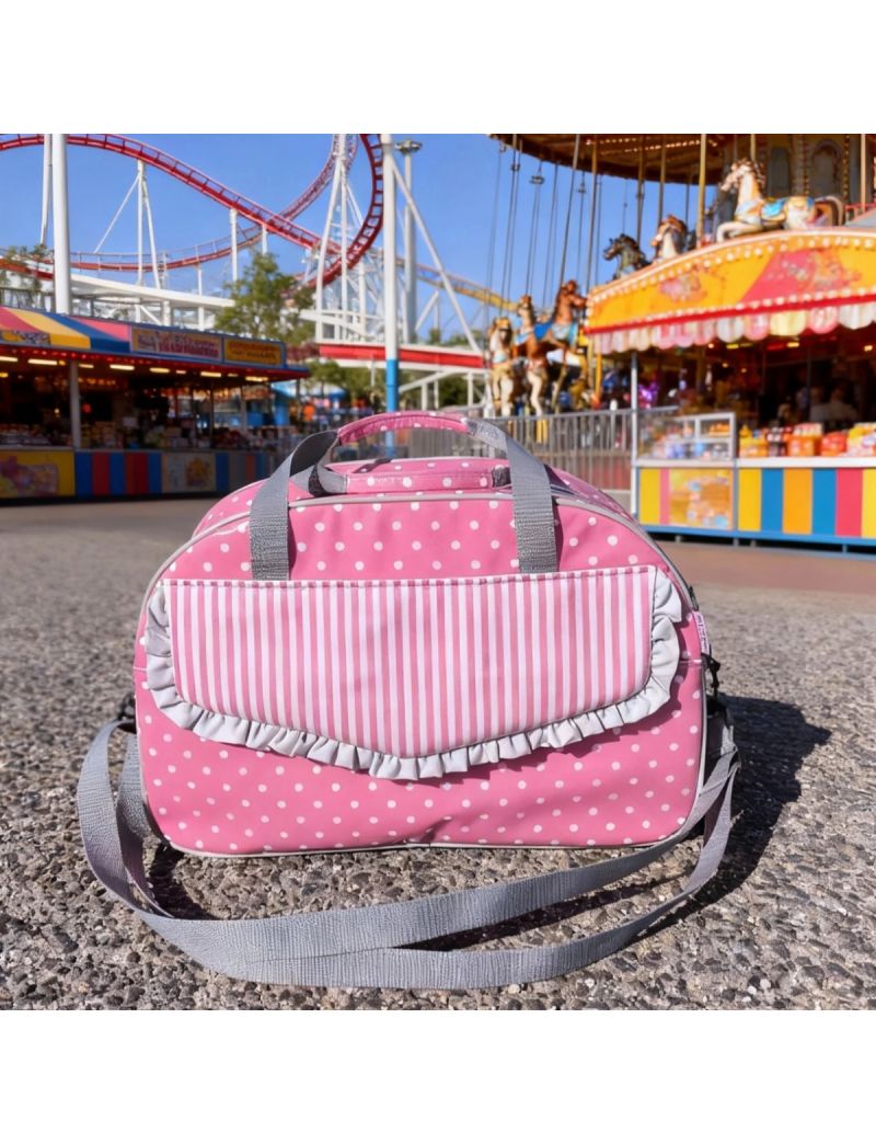 Saco de desporto cor-de-rosa com bolas brancas. Pala da bolsa frontal em risas rosa e brancas. Com folho na pala em cinzento. Alças em cinzento. Saco colocado um cerário de uma feira de diversões 