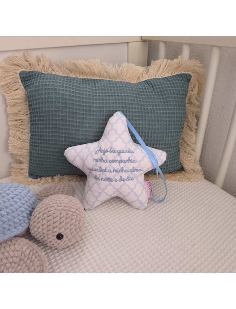 Estrela com a oração do Anjo da Guarda bordado a azul claro colocada numa caminha de bebé. Atrás tem uma almofada decorativa verde e ao lado uma tartaruga.