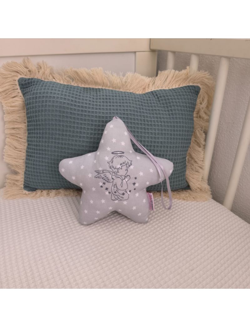 Estrela com um Anjo bordado a cinzento. A estrela é cinzenta com estrelas brancas. Colocada numa caminha de bebé, atrás tem uma almofada decorativa verde 