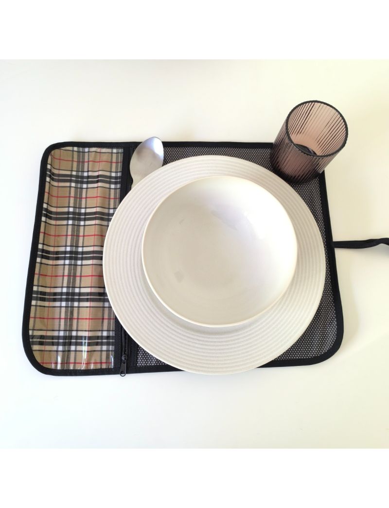Individual aberto com padrão de xadrez bege com riscas pretas na parte da bolsa para os talheres. Restante individual preto com bolinhas brancas. Prato, talheres e copo colocado no individual