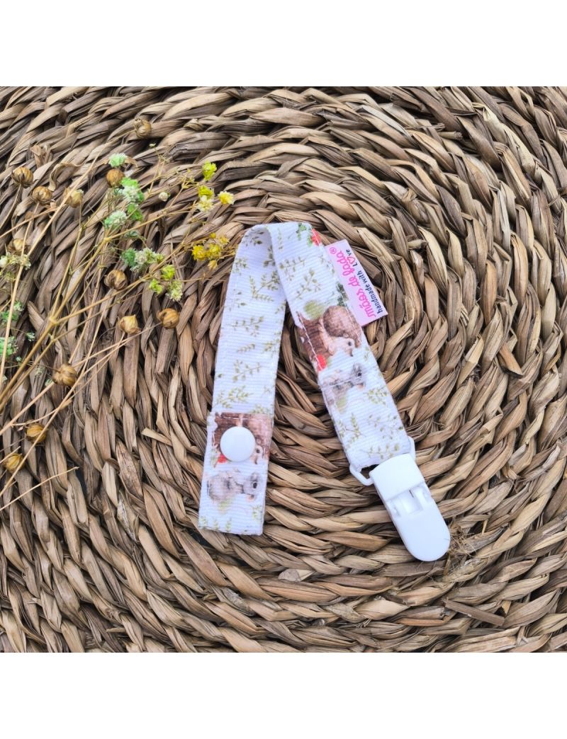 Fita de chucha em com padrão de animais e folhagens pequenas, com mola de plástico branca, colocada sobre base de palha artesanal. 
