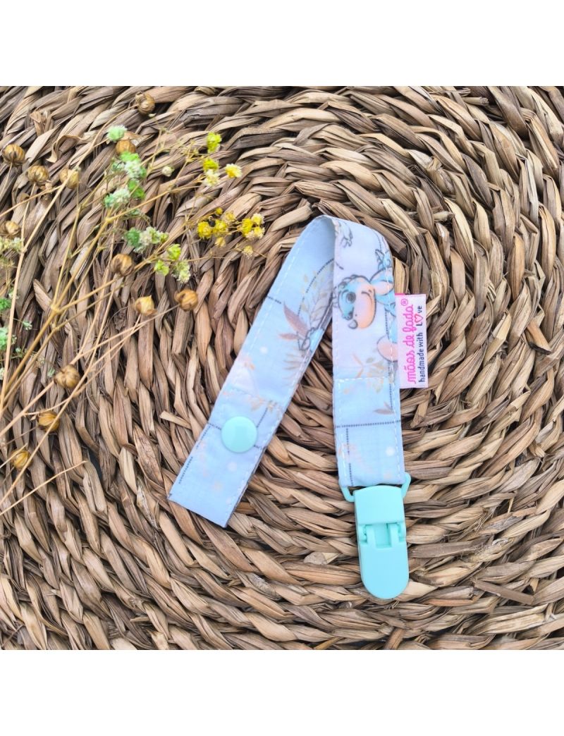 Fita de chucha com padrão de dinossauros em tons de verde água, com mola de plástico verde, colocada sobre base de palha artesanal. 