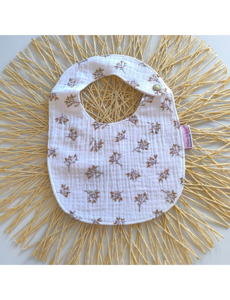 Babete de musselina branca com ramos em tons de camel, fecho de mola, impermeável, ideal para bebés até aos 7 meses. 
