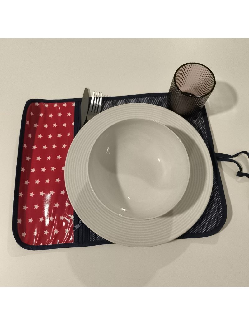 Individual aberto. Bolso para os talheres com tecido vermelho com strelas brancas e restante individual azul escuro com bolinhas brancas. Prato, talheres e copo colocado no individual