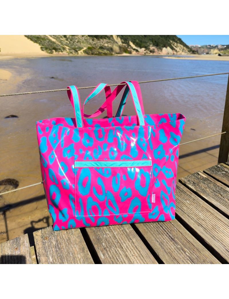 Saco de praia plastificado com padr&atilde;o de leopardo em tons rosa f&uacute;csia e verde &aacute;gua escuro. A mala tem um bolso exterior vis&iacute;vel e al&ccedil;as bicolores, estando pousada num passadi&ccedil;o de madeira com uma paisagem de rio, dunas e c&eacute;u azul ao fundo.