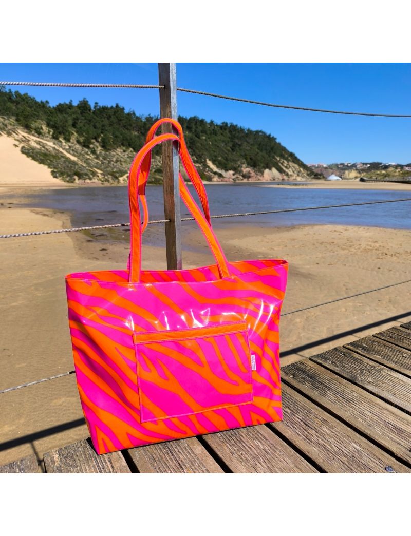 Saco de praia grande com padr&atilde;o zebra em tons n&eacute;on rosa e laranja vibrante. Em material plastificado imperme&aacute;vel o saco est&aacute; pendurado num passadi&ccedil;o de madeira com uma praia e dunas de fundo. Detalhe de bolso frontal e al&ccedil;as compridas no mesmo padr&atilde;o.