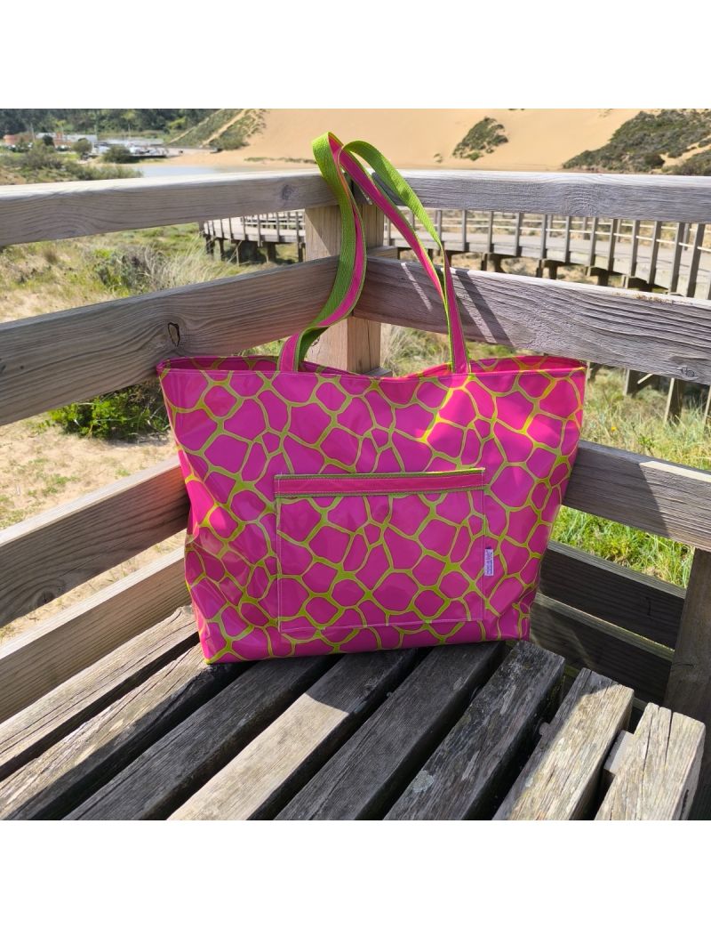 Saco de praia em tecido plastificado brilhante com padr&atilde;o que faz lembrar mosaico em rosa f&uacute;csia e linhas a verde lima. Possui um bolso frontal retangular e al&ccedil;as rosa e verdes, estando pousada num banco de madeira num passadi&ccedil;o com vista para dunas e nat
