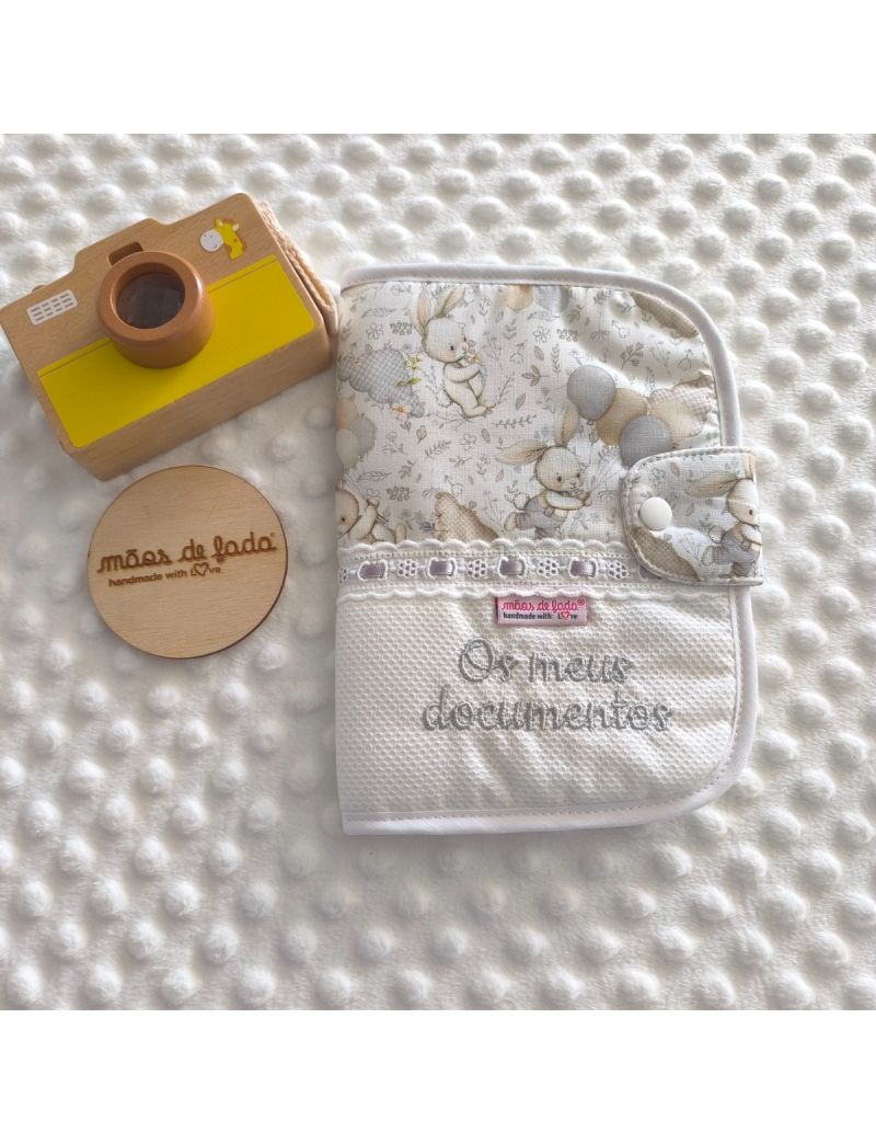 Porta documentos em formato livro com padr&atilde;o de coelhos e bal&otilde;es em tons neutros, com bordado ingl&ecirc;s com fita cetim cinza, com a inscri&ccedil;&atilde;o &ldquo;Os meus documentos&rdquo; bordado a cinza

