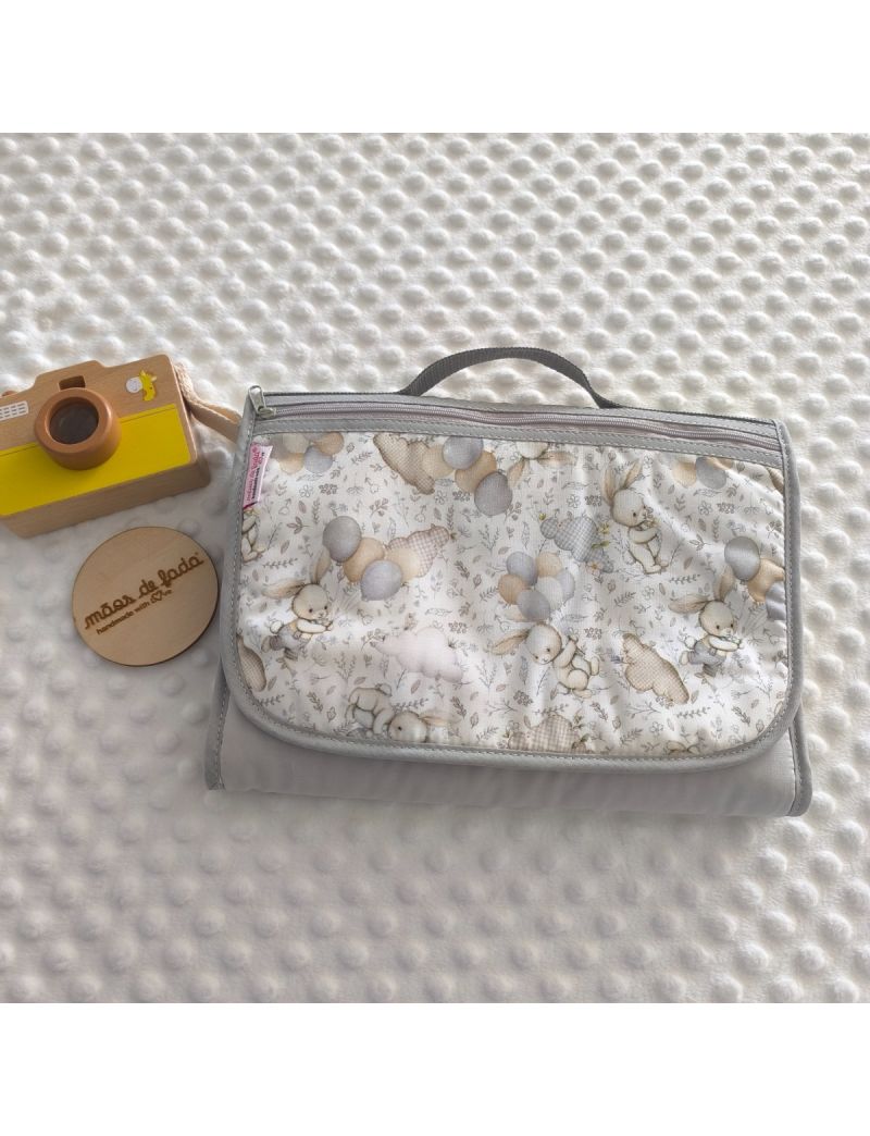 Bolsa multiusos para beb&eacute; fechada, com acabamentos em cinza claro e uma al&ccedil;a de m&atilde;o curta na parte superior. O tecido exterior apresenta um padr&atilde;o delicado e suave de coelhinhos ilustrados com bal&otilde;es em tons pastel.