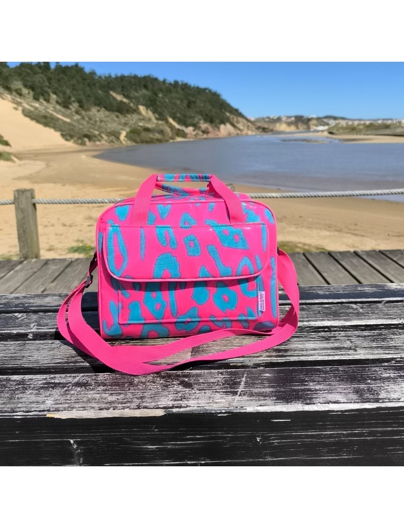 Lancheira t&eacute;rmica da cole&ccedil;&atilde;o Leopard Neon com padr&atilde;o animal em rosa choque e verde &aacute;gua escuro, pousada num banco de madeira com uma praia e dunas ao fundo. A lancheira apresenta al&ccedil;as de m&atilde;o e uma al&ccedil;a a tiracolo ajust&aacute;vel em rosa.