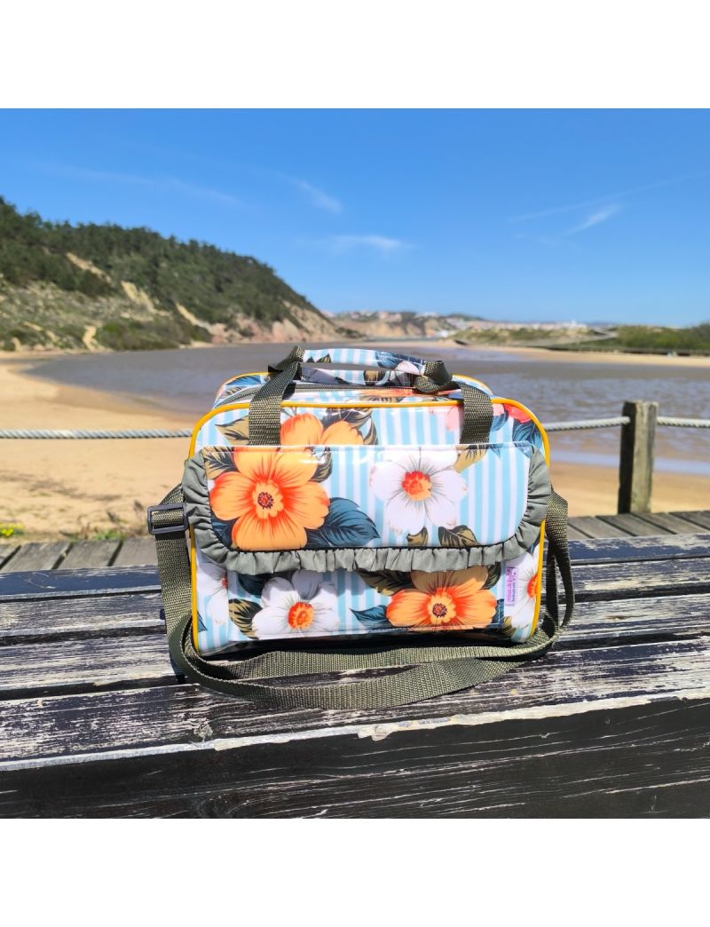 Lancheira t&eacute;rmica com padr&atilde;o floral tropical (flores laranja e brancas sobre riscas) pousada num banco de madeira r&uacute;stico. A lancheira tem um detalhe de folho verde seco na bolsa frontal, al&ccedil;a de m&atilde;o e al&ccedil;a a tiracolo regul&aacute;vel em verde