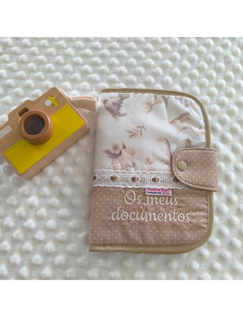 Porta documentos em formato livro com tecido creme com bolinhas brancas conjugado com um padr&atilde;o de dinossauros em tons suaves. Possui bordado ingl&ecirc;s com fita de cetim bege e a inscri&ccedil;&atilde;o "Os meus documentos"