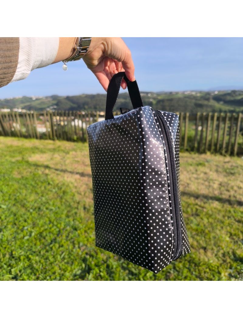 Uma pessoa segura, pela al&ccedil;a superior (preta), uma necessaire retangular com fundo preto e bolinhas brancas