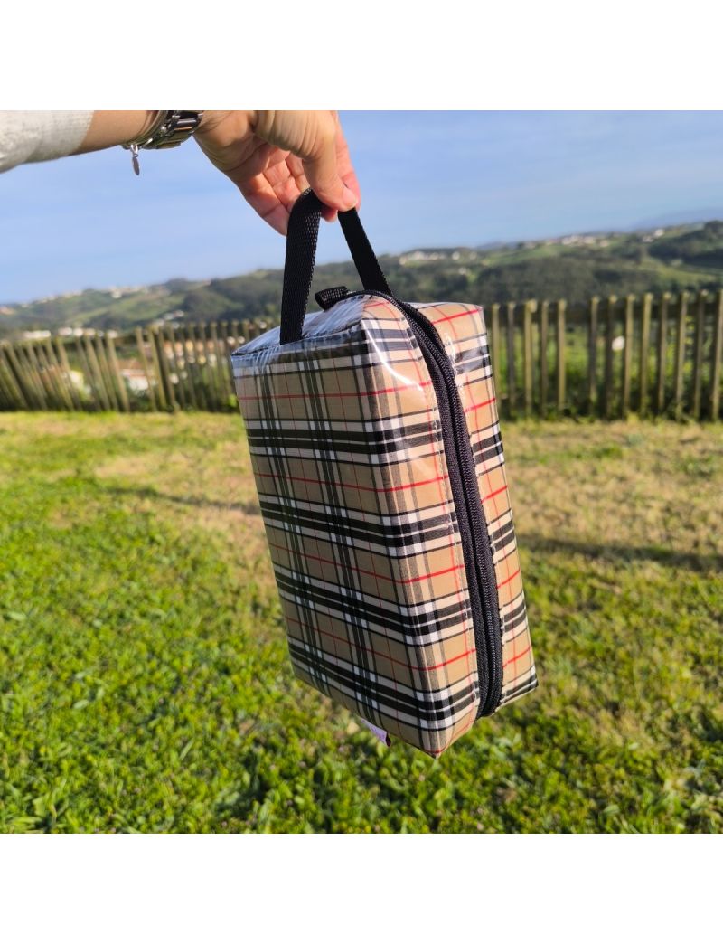 Necessaire box  em tecido plastificado bege, com padr&atilde;o xadrez cl&aacute;ssico em tons de preto, branco e vermelho, segurada pela al&ccedil;a de m&atilde;o preta