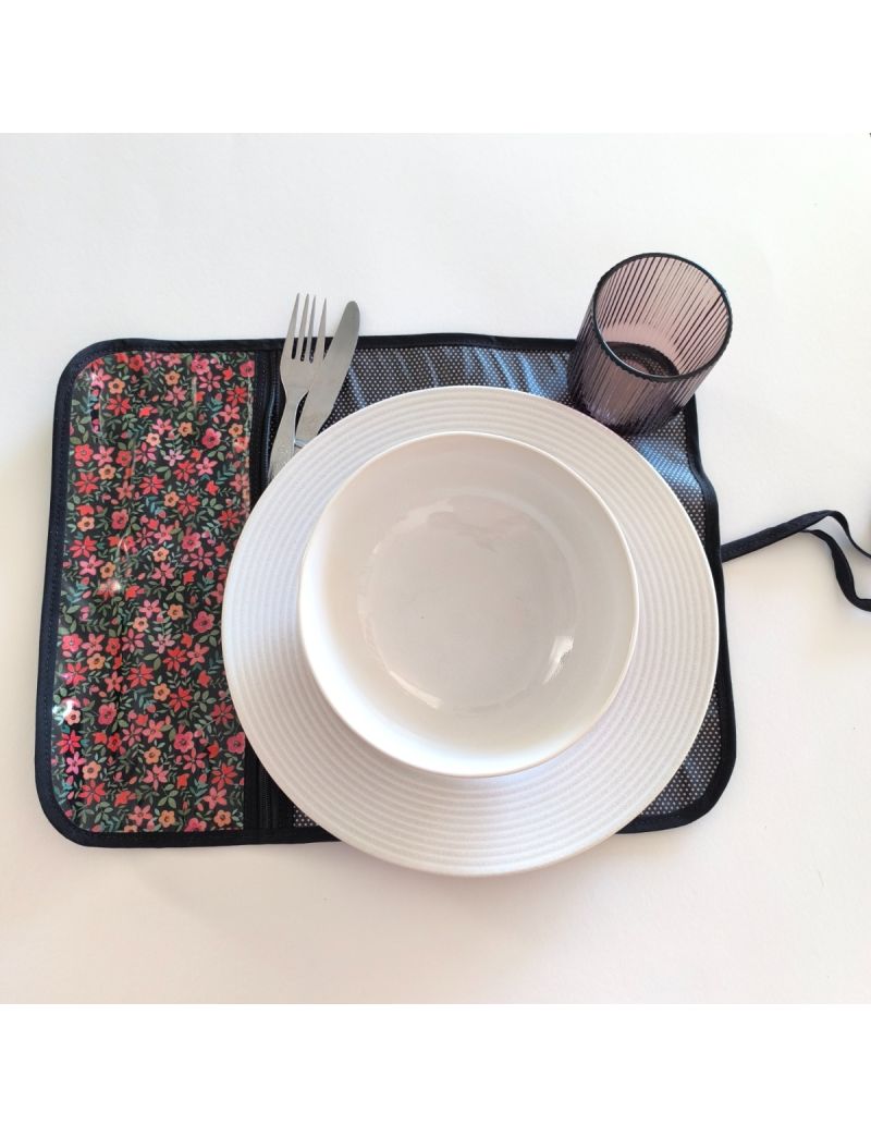 Individual aberto. Bolso para os talheres em tecido preto com florzinhas em tons de salm&atilde;o, restante individual preto com bolinhas brancas. Prato, talheres e copo colocado no individual