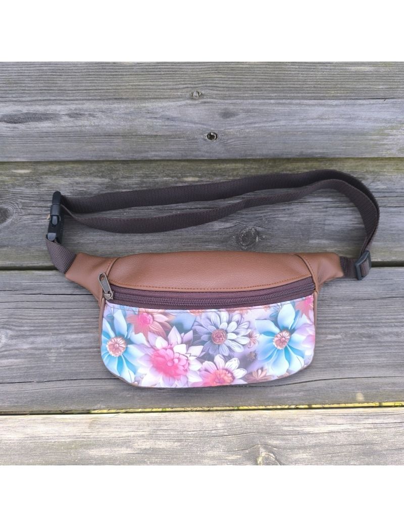 Bolsa de cintura em material sint&eacute;tico castanho-cl&aacute;ssico com um painel frontal em padr&atilde;o floral vibrante e art&iacute;stico em tons de azul e rosa. A bolsa possui um fecho de correr castanho, al&ccedil;a castanha ajust&aacute;vel com fecho de encaixe r&aacute;pido e est&aacute; pousada sob