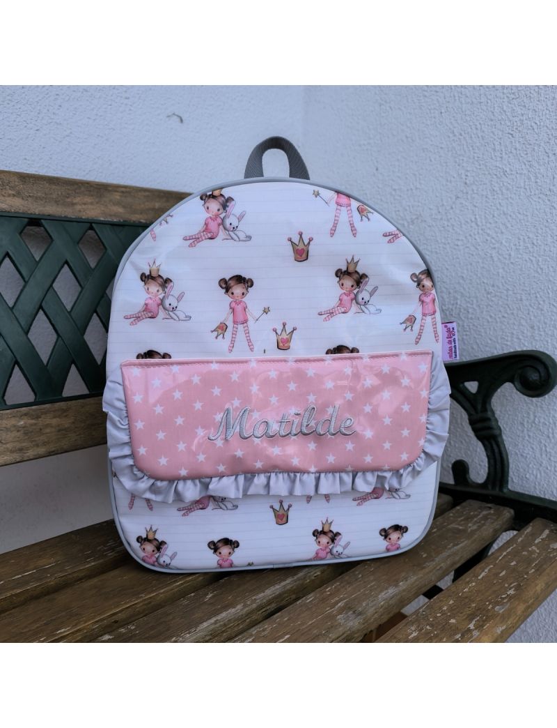 Mochila de criança com bonequinhas. Bolsa frontal com pala com tecido rosa pastel com estrelas brancas e o nome Matilde bordado a cinzento. Folho na pala cinzento.