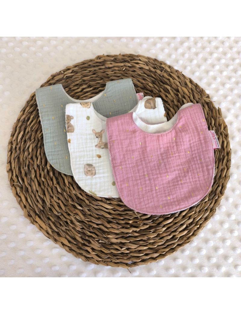 Conjunto de tr&ecirc;s babetes de mola em musselina. Inclui um babete verde, um babete branco central com ilustra&ccedil;&otilde;es de animais da floresta e um babete rosa velho. Todos apresentam pequenos pontos dourados e est&atilde;o dispostos de forma sobreposta numa base de ver