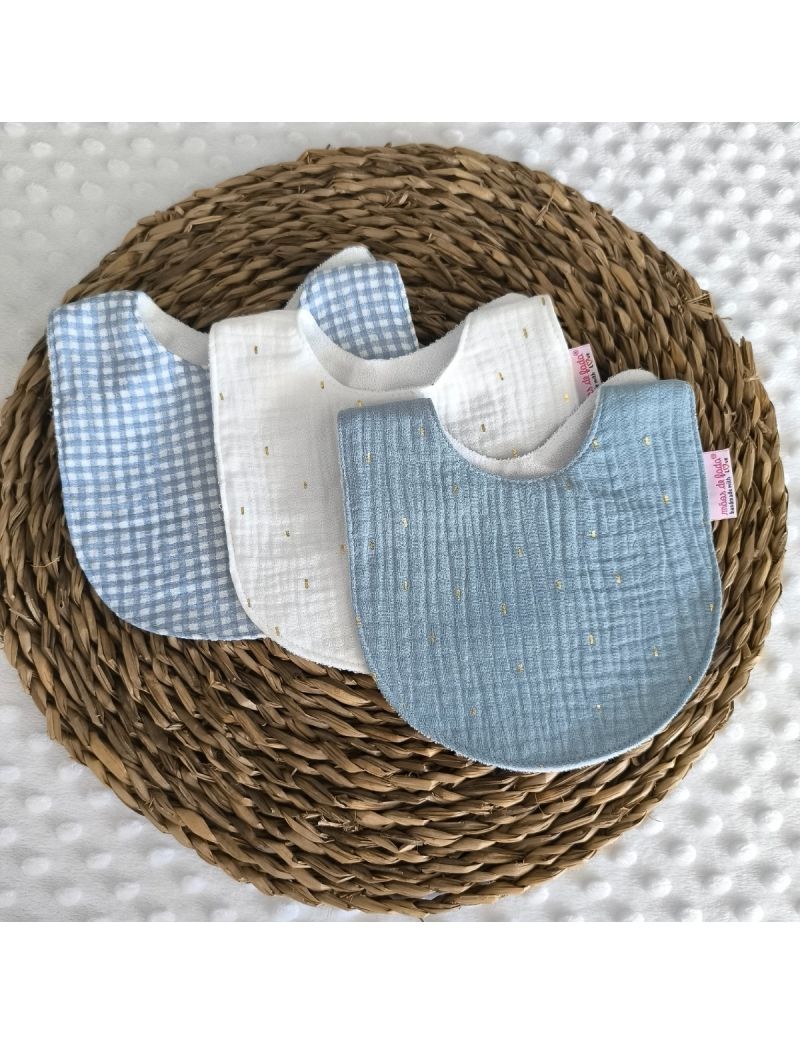 Conjunto de tr&ecirc;s babetes de mola em musselina dispostos numa base de verga. &Agrave; frente, um babete azul com pequenos pontos dourados; ao centro, um babete branco com o mesmo detalhe; atr&aacute;s, um babete com padr&atilde;o aos quadrados (vichy) em azul e branco