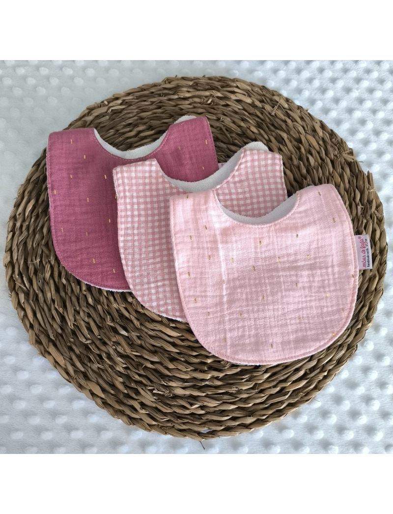 Conjunto de tr&ecirc;s babetes redondos de mola em musselina de algod&atilde;o dispostos sobre uma base de verga. &Agrave; frente, um babete rosa claro com pequenos pontos dourados; ao centro, um babete com padr&atilde;o xadrez vichy rosa e branco; atr&aacute;s, um babete em tom amora esc