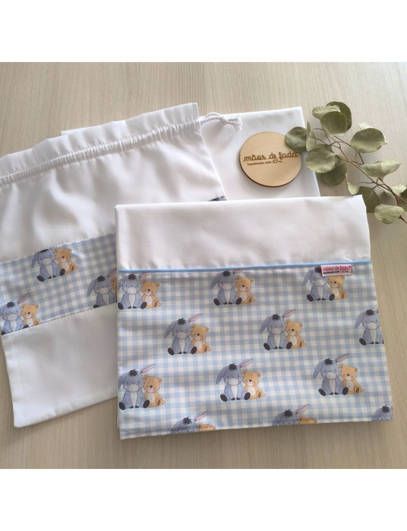 Lençol de cima, lençol de baixo e saquinho. O lençol de cima e o saquinho possuem uma barra com padrão de quadradinhos azuis e brancos com o burro e o urso do Winnie de Pooh