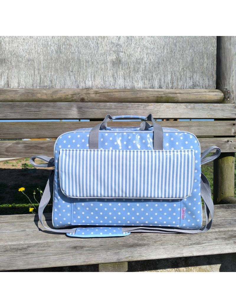 Bolsa de maternidade azul com padrão de bolas brancas. Pala frontal com riscas azul e brancas. Alça de ombro ajustável em cinzento. Mala colocada sobre um banco de madeira ao ar livre