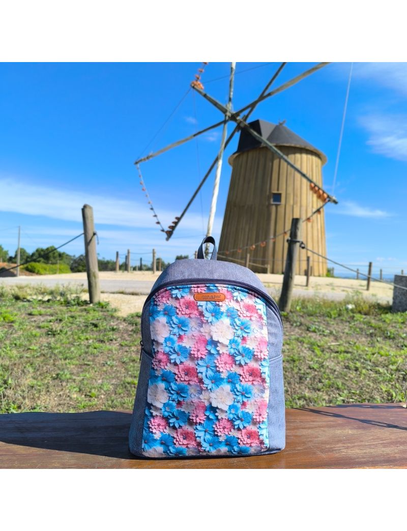 Mochila de senhora artesanal azul ganga com painel frontal em tecido estampado de flores coloridas. Pousada em madeira, com moinho português em fundo