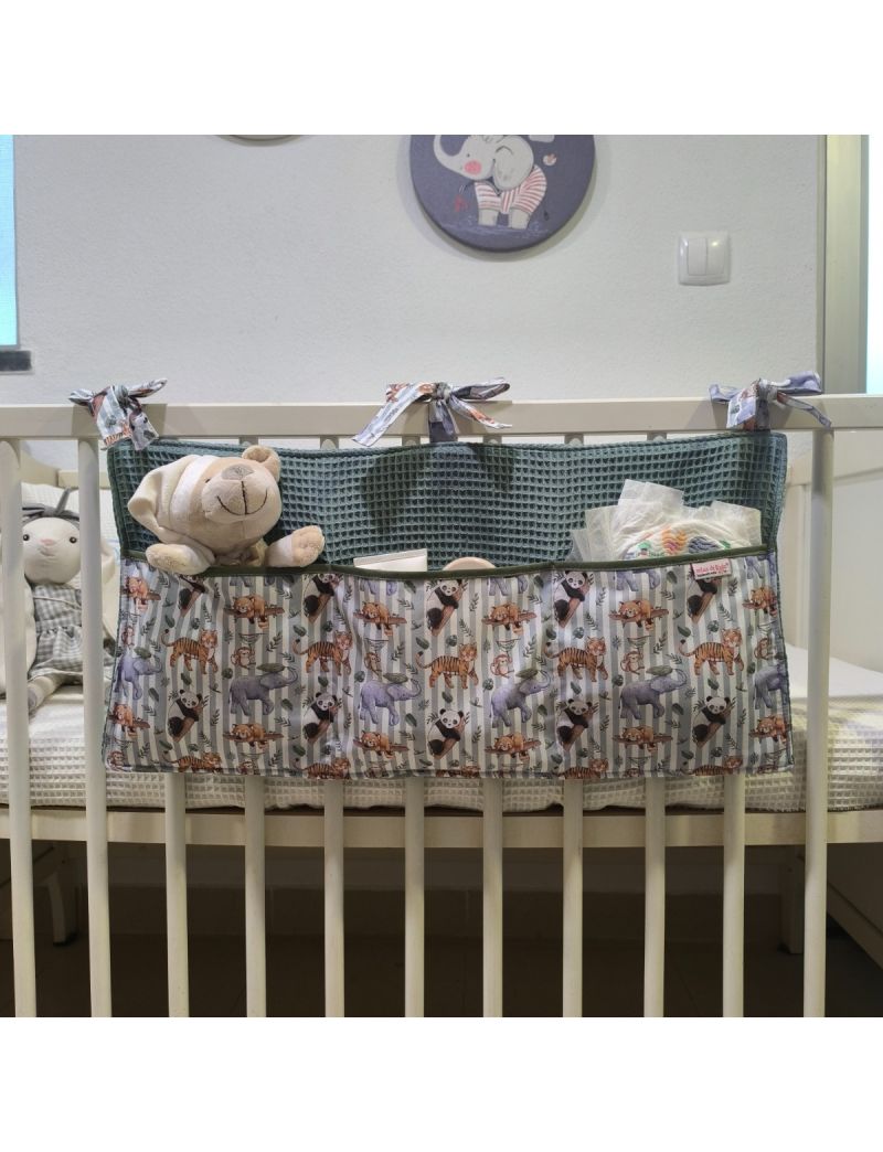 Organizador de cama para bebé em tecido com animais conjugado com waffle verde, com três bolsos, pendurado num berço branco e com brinquedos e acessórios de bebé no interior