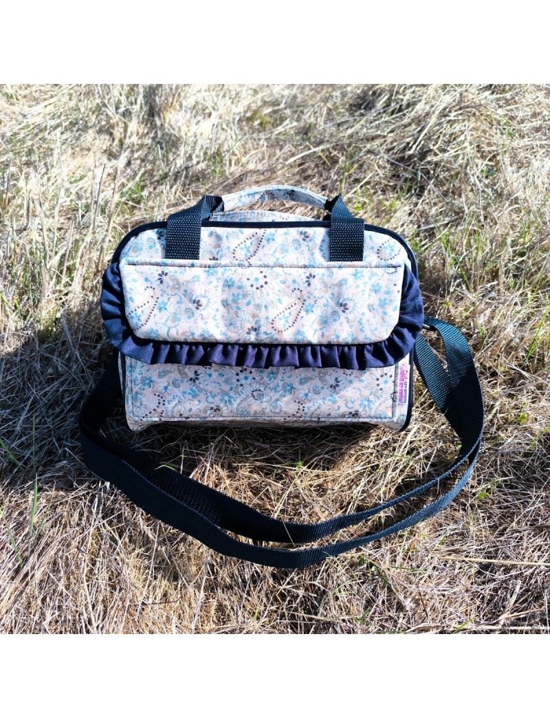 Lancheira XS com alça ajustável e alça de mão azul escura, com padrão floral em tons de azul, e detalhe de folho em azul escuro na pala frontal, colocada sobre relva seca ao ar livre