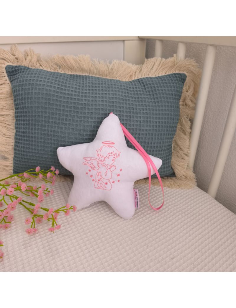 Estrela branca com anjo bordado a rosa, fita rosa e enchimento antialérgico, apoiada em almofadas decorativas num quarto de bebé.