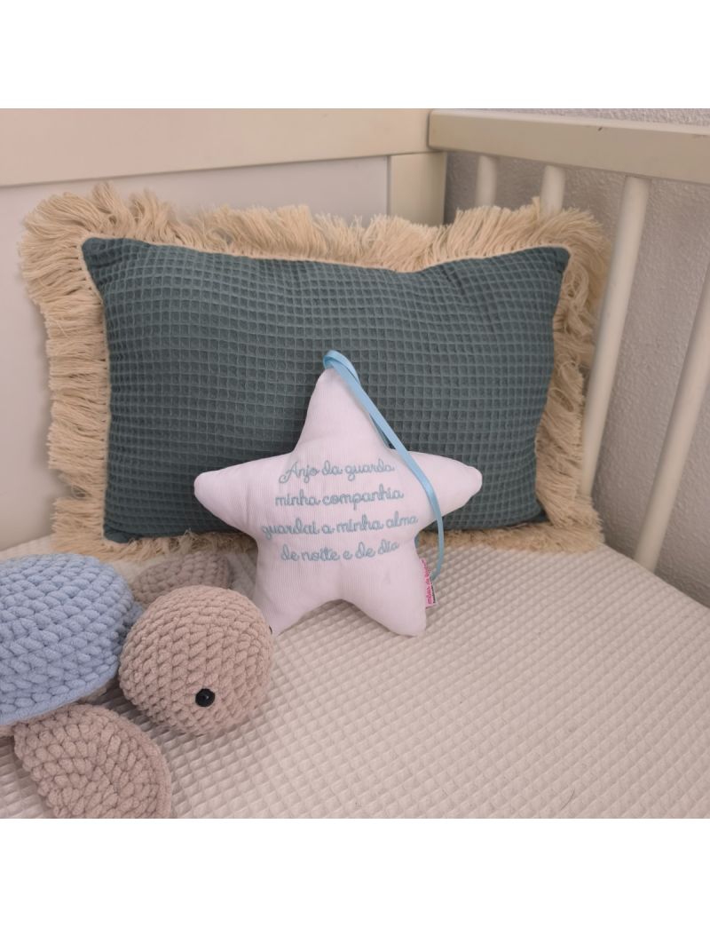 Estrela com a oração do Anjo da Guarda bordado a azul claro colocada numa caminha de bebé. Atrás tem uma almofada decorativa verde e ao lado uma tartaruga