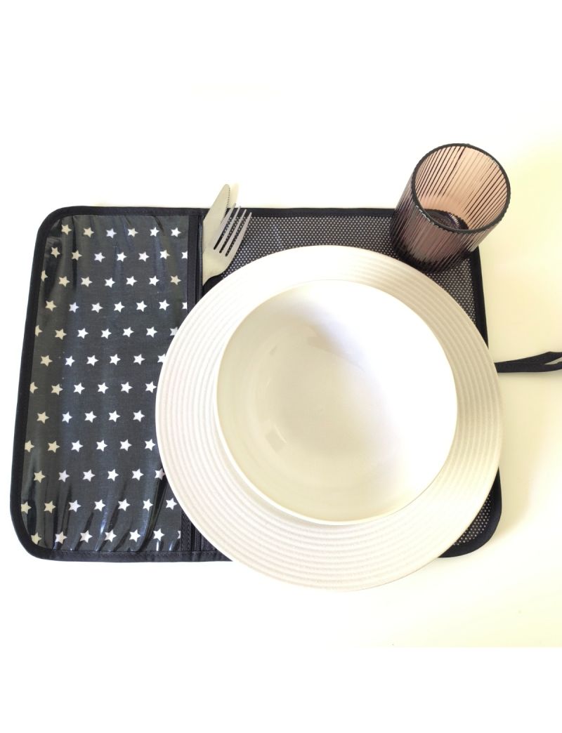 Individual aberto preto com estrelas brancas na parte da bolsa para os talheres. Restante individual preto com bolinhas brancas. Prato, talheres e copo colocado no individual