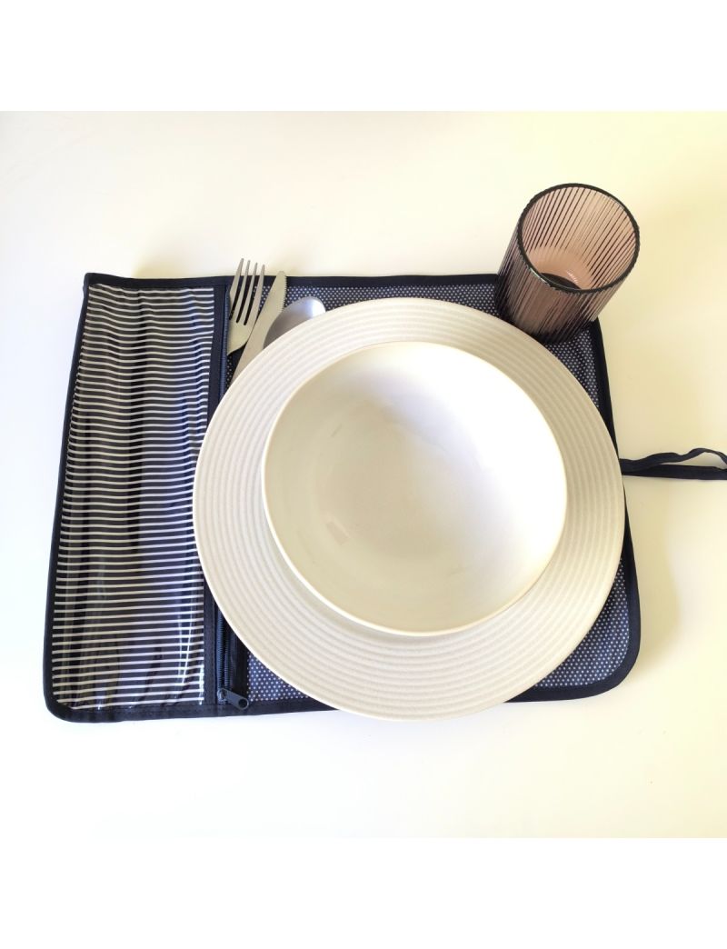 Individual aberto com padrão de riscas azuis escuras e brancas na parte da bolsa para os talheres. Restante individual azul escuro com bolinhas brancas. Prato, talheres e copo colocado no individual
