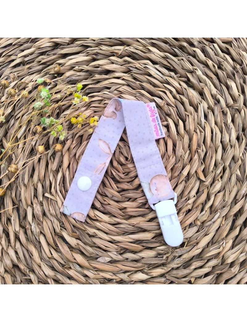 Fita de chucha com padrão de ursinhos em tons de bege e castanho, com mola de plástico branca, colocada sobre base de palha artesanal. 