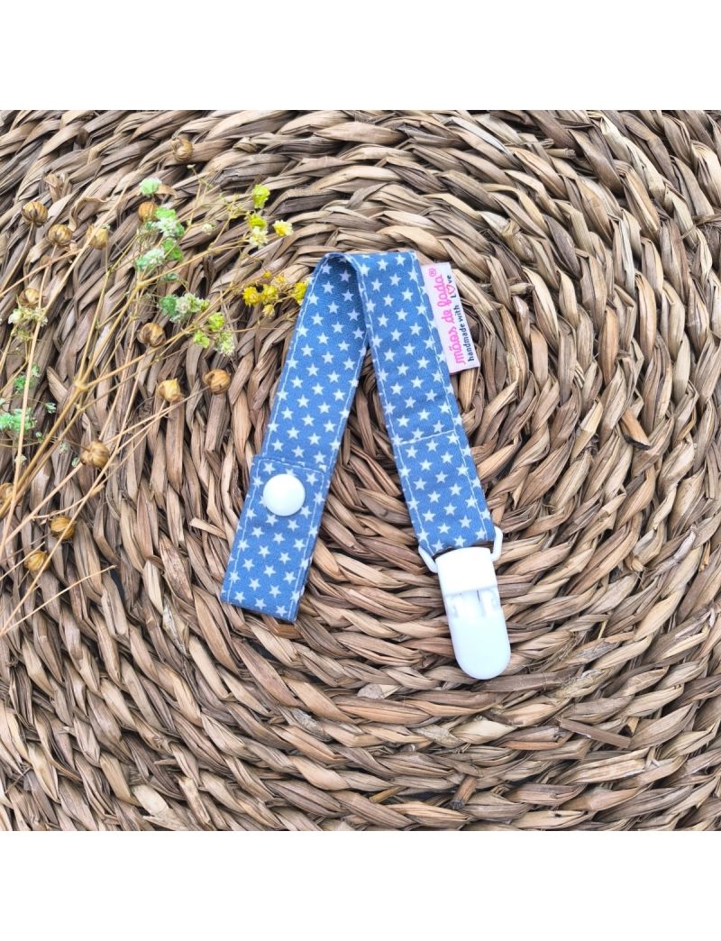 Fita de chucha em tecido de algodão azul com estrelinhas, com mola de plástico branca, colocada sobre base de palha artesanal.