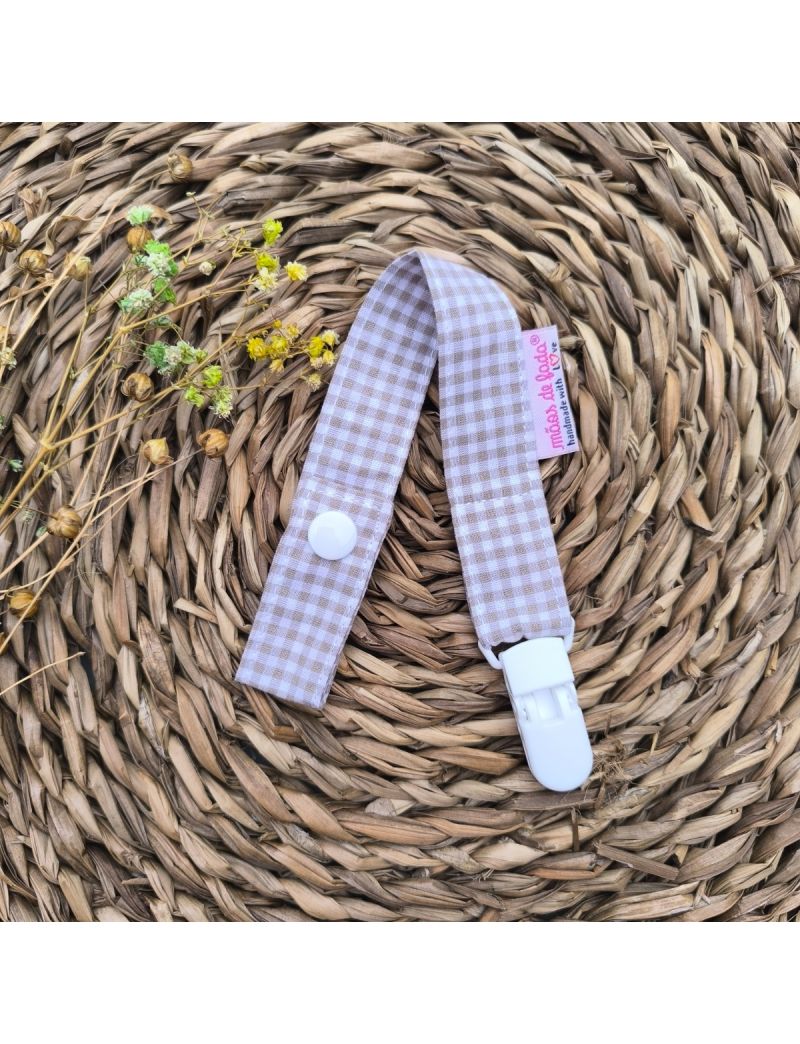 Fita de chucha em tecido de algodão xadrez vichy castanho, com mola de plástico branca, colocada sobre base de palha artesanal. 