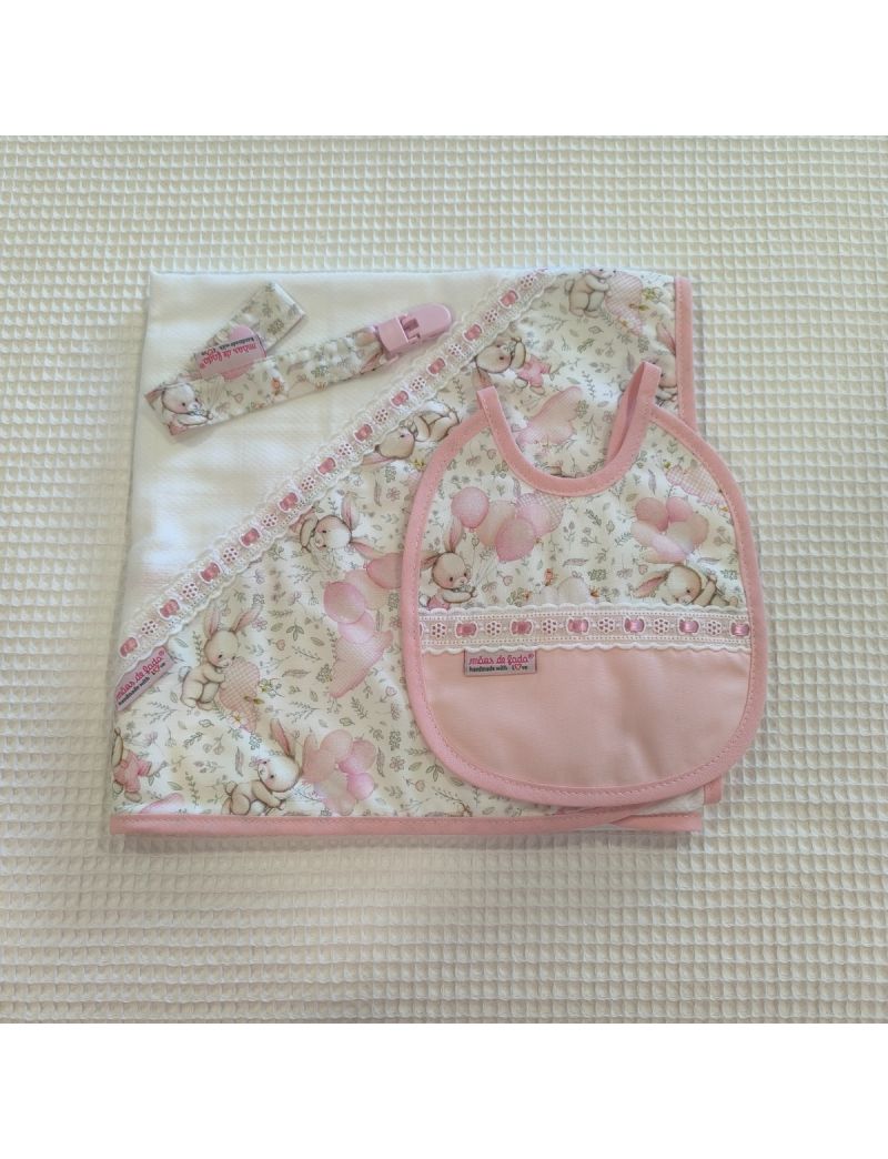 Conjunto de bebé composto por fralda branca com triângulo de tecido com padrão de coelhos e balões em tons de cor de rosa. Babete com padrão igual à fralda conjugado com tecido rosa liso e com bordado inglês com fita de cetim rosa. Fita para a chucha com 