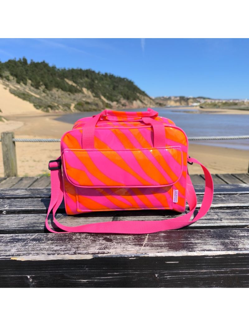 Lancheira t&eacute;rmica da cole&ccedil;&atilde;o Neon  Zebra com padr&atilde;o de riscas de zebra em rosa choque e laranja vibrante. Est&aacute; pousada num banco de madeira com fundo de praia, destacando a bolsa frontal e as al&ccedil;as de m&atilde;o e tiracolo em rosa.