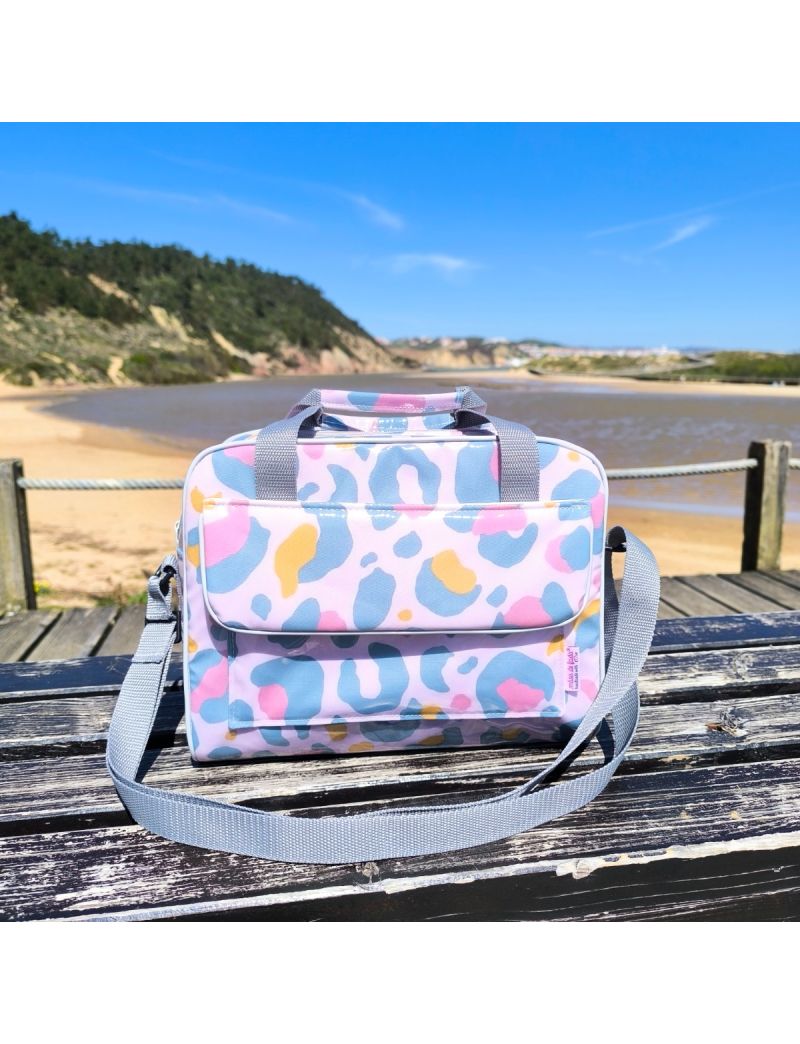 Lancheira t&eacute;rmica com padr&atilde;o de leopardo em tons de rosa, amarelo e verde &aacute;gua escuro pousada num banco de madeira r&uacute;stico. A lancheira tem uma bolsa frontal com pala, al&ccedil;as de m&atilde;o e uma al&ccedil;a a tiracolo ajust&aacute;vel em cinza.