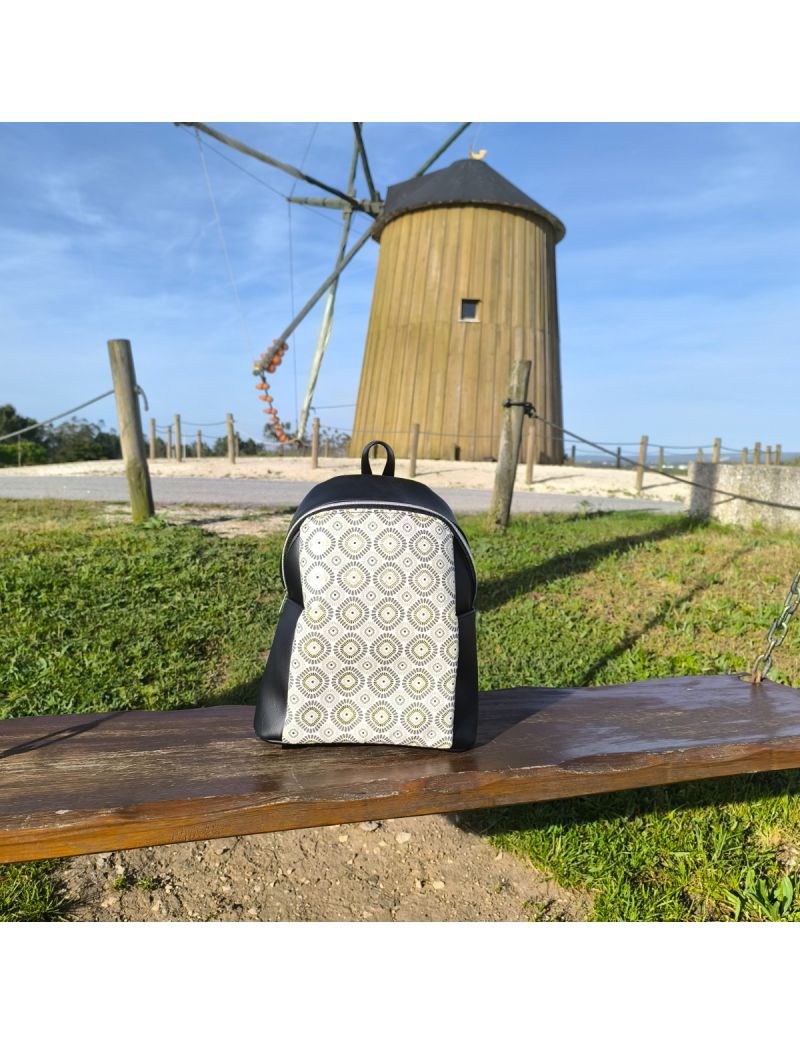 Mochila de senhora em sint&eacute;tico preto com painel frontal em tecido de padr&atilde;o geom&eacute;trico bege e cinzento, pousada num banco de madeira com um moinho de vento ao fundo.