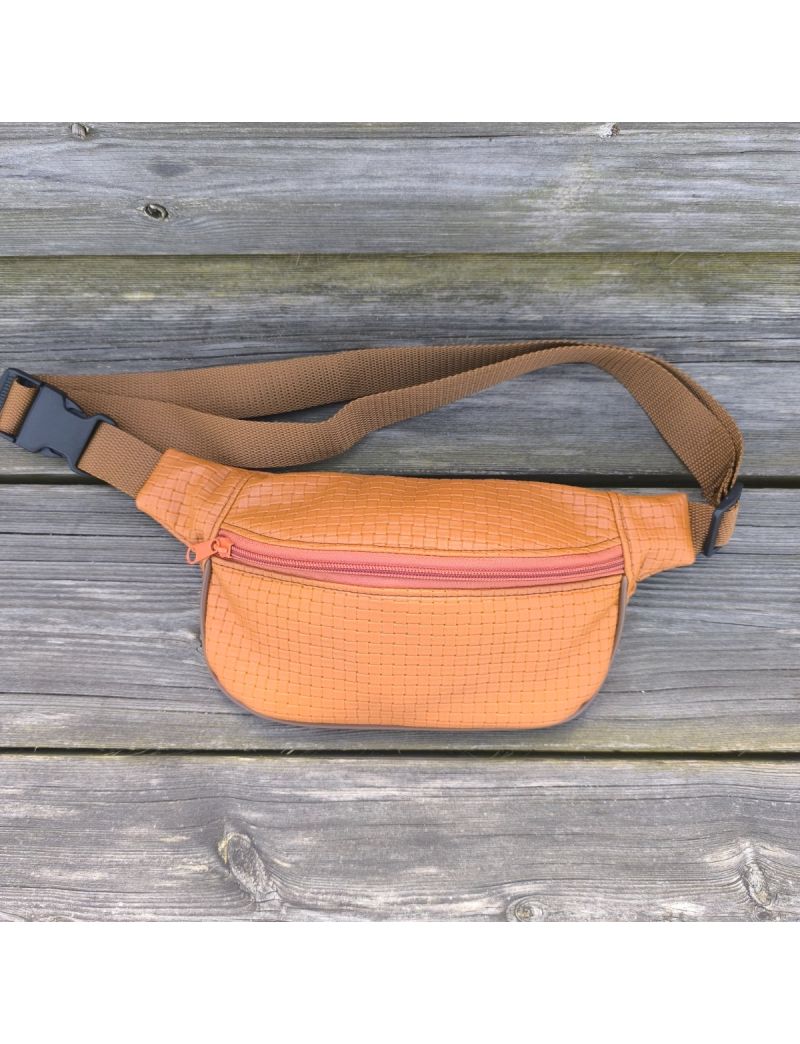 Bolsa de cintura em material sint&eacute;tico cor terracota com textura de padr&atilde;o entran&ccedil;ado. Possui fecho de correr na mesma cor, al&ccedil;a castanha ajust&aacute;vel com fivela de encaixe preta e est&aacute; pousada sobre um banco de madeira r&uacute;stica 