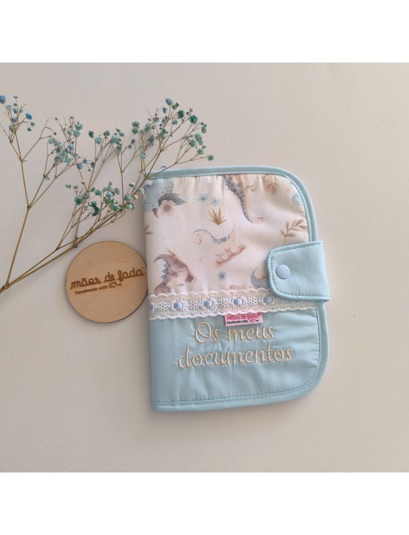 Porta documentos em formato livro com tecido azul conjugado com um padrão de dinossauros em tons suaves. Possui bordado inglês com fita de cetim azul e a inscrição "Os meus documentos"