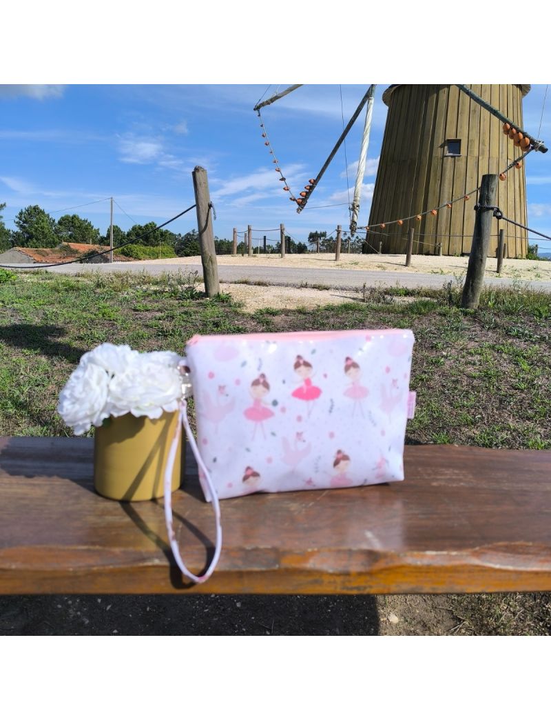 Necessaire em tecido plastificado com padrão de bailarinas, pousada sobre uma superfície de madeira, com fundo exterior iluminado e ambiente sereno